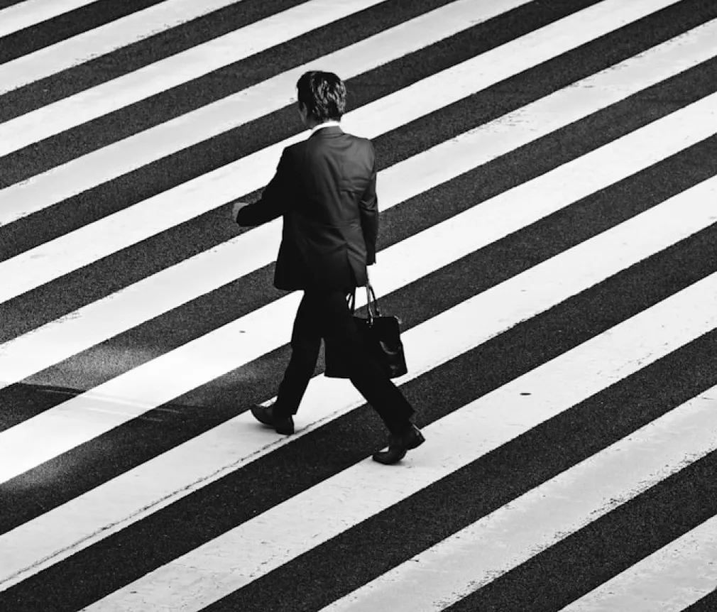 Black-and-white editorial photo of pedestrians crossing a busy city street